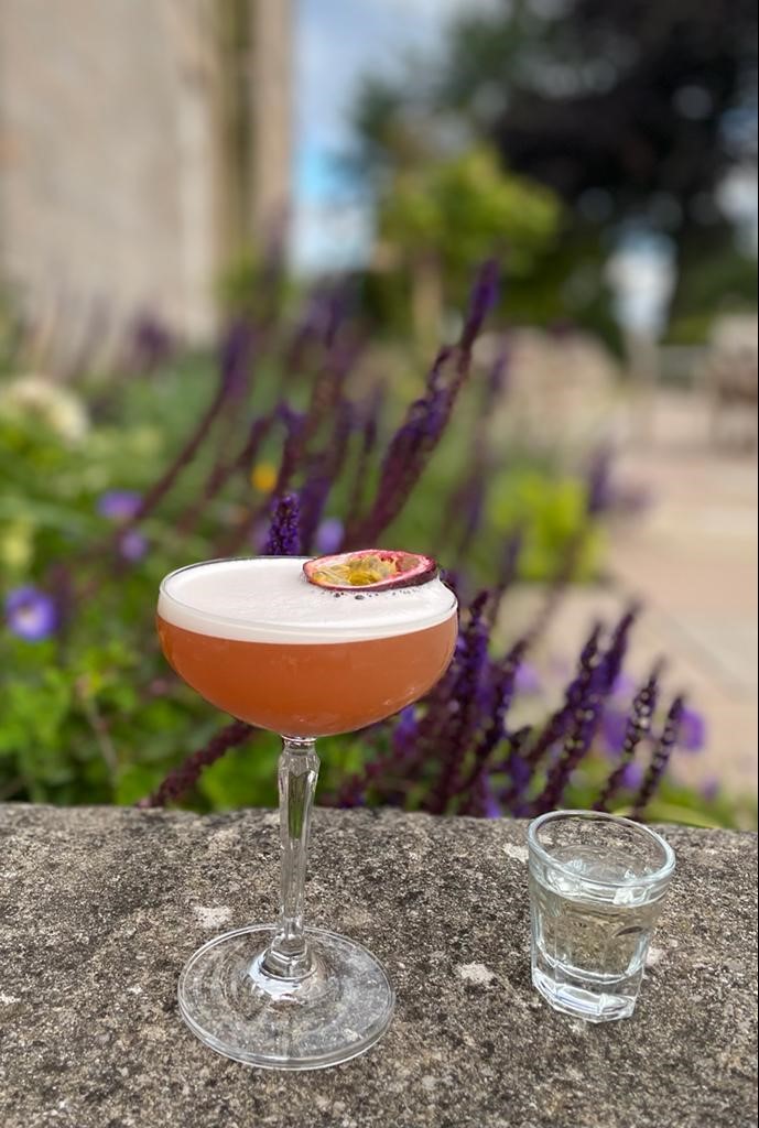Summer Breeze An orange-coloured cocktail in a cocktail glass with a slice of passionfruit placed on the top, next to a clear shot in a shot glass, perched on a ledge in the Marshall Meadows patio area